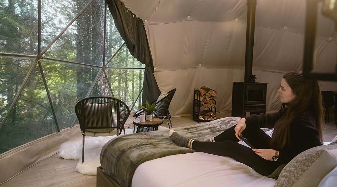 Interior of a forest-view geodesic dome with woman sitting on a bed overlooking the forest from a large window.