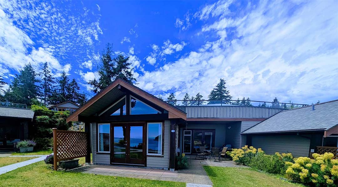 Exterior view of Mayne Island Resort featuring a lodge-style building, landscaped grounds, and a bright blue sky with mountains in the background.